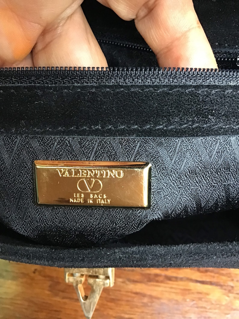 May include: Close-up of a black handbag interior with a gold-colored metal plaque. The plaque reads "VALENTINO LES SACS MADE IN ITALY". The bag has a zipper and a gold-colored clasp.
