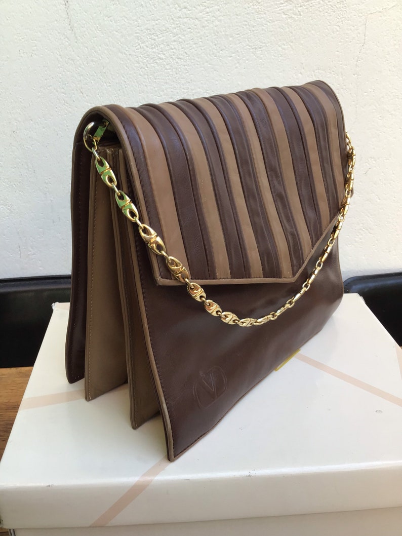 May include: A brown and tan leather handbag with a gold chain strap. The flap features vertical pleats, and the bag has a gold chain strap. The Valentino logo is embossed on the front.