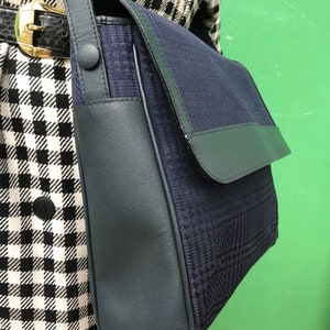 May include: A navy blue shoulder bag with a flap closure. The bag features a textured, patterned fabric body and dark blue leather trim. The strap is also dark blue, and the bag is secured with a button closure.