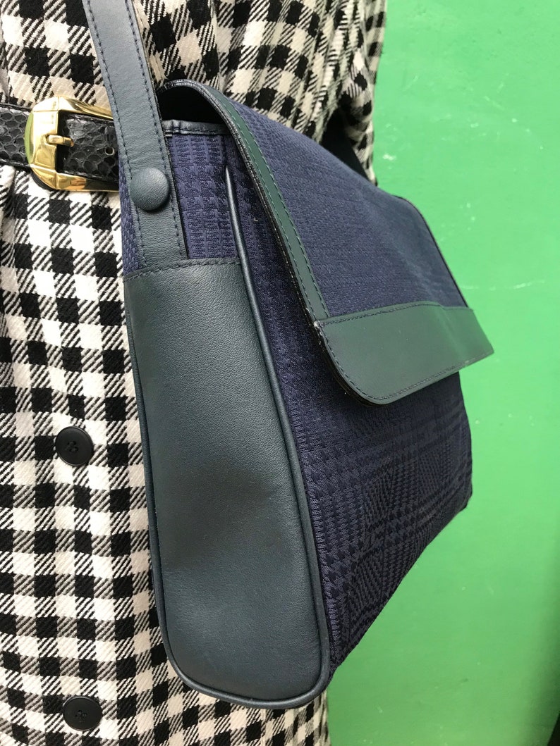 May include: A navy blue shoulder bag with a flap closure. The bag features a textured, patterned fabric body and dark blue leather trim. The strap is also dark blue, and the bag is secured with a button closure.