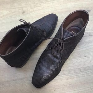 Può includere: Un paio di stivaletti alla caviglia in pelle marrone scuro con un motivo strutturato simile alla pelle di serpente. Gli stivaletti hanno lacci e un tacco basso. L'interno degli stivaletti è di colore marrone chiaro.