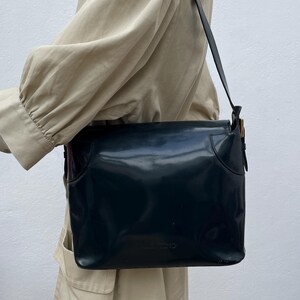 Puede incluir: Un bolso de hombro azul marino oscuro con un acabado liso y brillante y un cierre de solapa rectangular. El bolso cuenta con una correa de hombro &uacute;nica y detalles de herrajes en tono dorado en el lateral. El nombre Valentino est&aacute; grabado al frente.