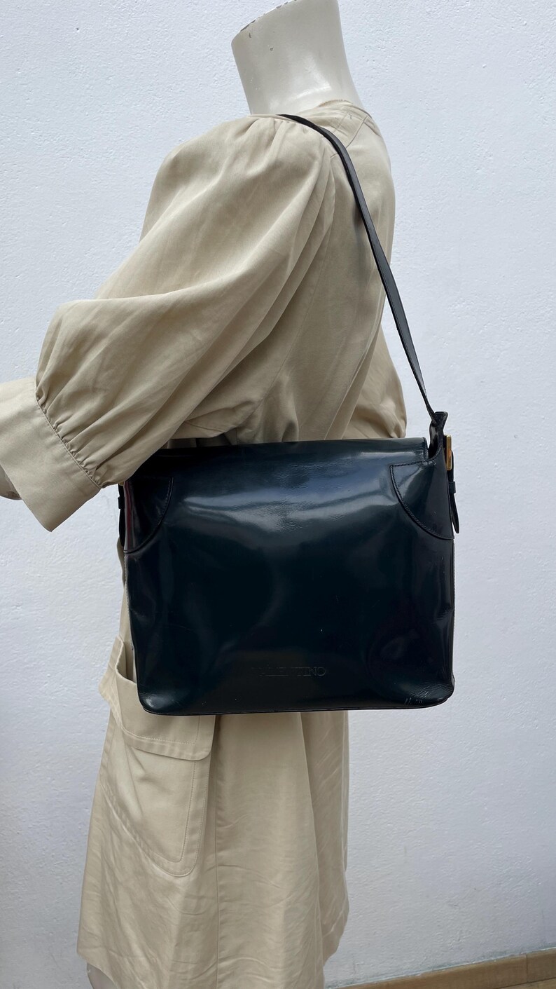 Puede incluir: Un bolso de hombro azul marino oscuro con un acabado liso y brillante y un cierre de solapa rectangular. El bolso cuenta con una correa de hombro &uacute;nica y detalles de herrajes en tono dorado en el lateral. El nombre Valentino est&aacute; grabado al frente.