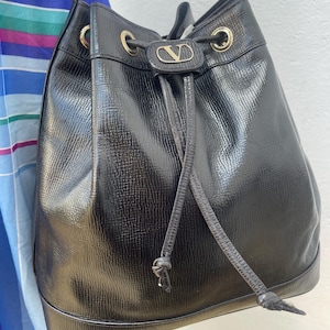 May include: Black leather bucket bag with a drawstring closure and a shoulder strap. The bag features gold-tone hardware and a small logo emblem on the front. The bag has a textured surface.