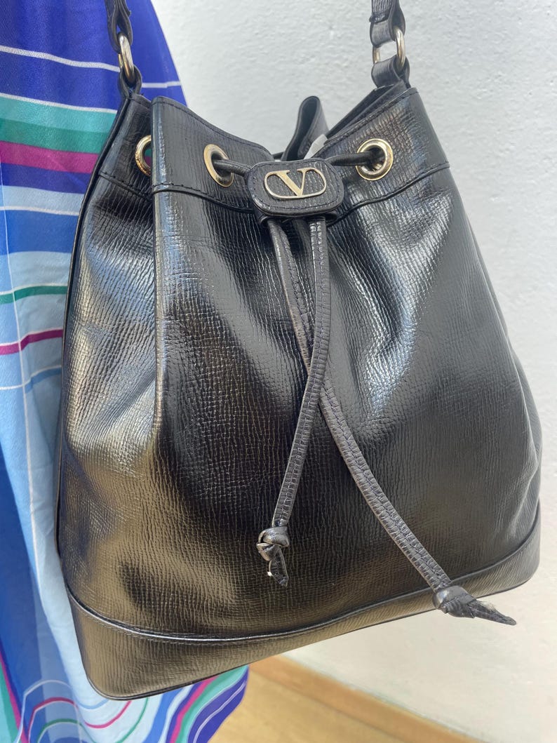 May include: Black leather bucket bag with a drawstring closure and a shoulder strap. The bag features gold-tone hardware and a small logo emblem on the front. The bag has a textured surface.
