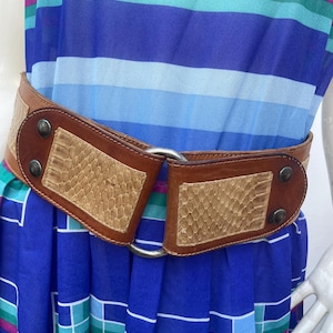 Può includere: Una cintura in pelle marrone con motivo a pelle di serpente e fibbia color argento. La cintura è larga e presenta un pannello rettangolare in pelle di serpente su ciascun lato. La cintura è indossata sopra un vestito blu a righe orizzontali.