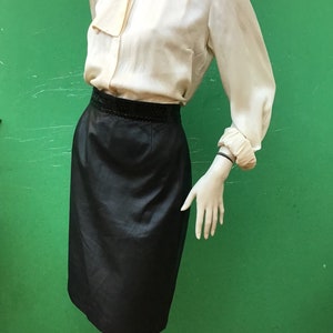 Puede incluir: Una blusa color crema con lazo en el cuello y mangas largas, combinada con una falda negra hasta la rodilla. La falda tiene una banda decorativa en la cintura. El conjunto se exhibe en un maniquí sobre un fondo verde.