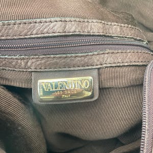 May include: Close-up of a brown leather handbag interior. The bag features a zipper and a metal tag that reads "VALENTINO LES SACS ITALY". The interior lining has a textured, striped pattern.