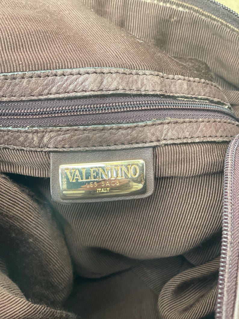 May include: Close-up of a brown leather handbag interior. The bag features a zipper and a metal tag that reads "VALENTINO LES SACS ITALY". The interior lining has a textured, striped pattern.