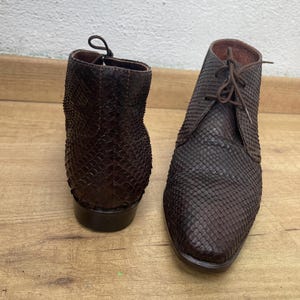 Puede incluir: Un par de botines de cuero marrón con un patrón texturizado en forma de escamas. Las botas tienen cordones y un tacón bajo. Una bota está orientada hacia la cámara, mientras que la otra está de espaldas.