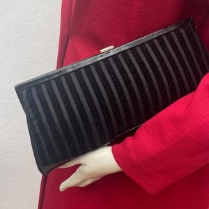 Può includere: Una pochette nera con strisce verticali in velluto e una chiusura color oro. La borsa rettangolare è tenuta contro un capo rosso, creando un contrasto sorprendente. La pochette è un accessorio classico.