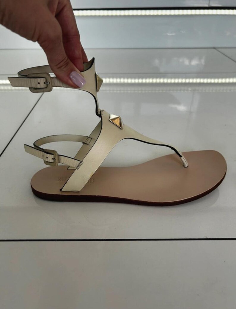 May include: Cream-colored leather sandals with a thong-style toe and ankle strap. The sandals feature gold-colored pyramid studs on the straps and a buckle closure. The sole is a light tan color.
