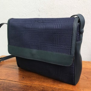 May include: A navy blue crossbody bag with a flap closure. The bag features a plaid pattern on the front and a dark green leather trim. The strap is adjustable and made of black leather.