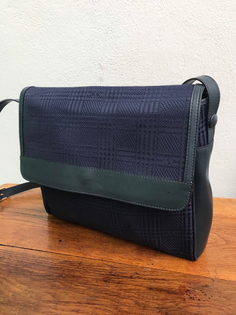 May include: A navy blue crossbody bag with a flap closure. The bag features a plaid pattern on the front and a dark green leather trim. The strap is adjustable and made of black leather.