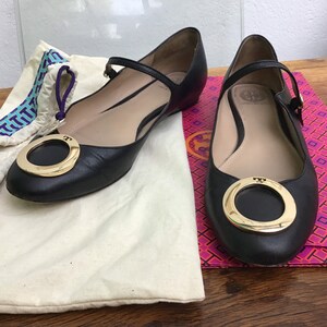 May include: Black leather ballet flats with a gold-tone ring detail on the toe. The flats have a strap across the top of the foot and are displayed with a dust bag and a patterned box.