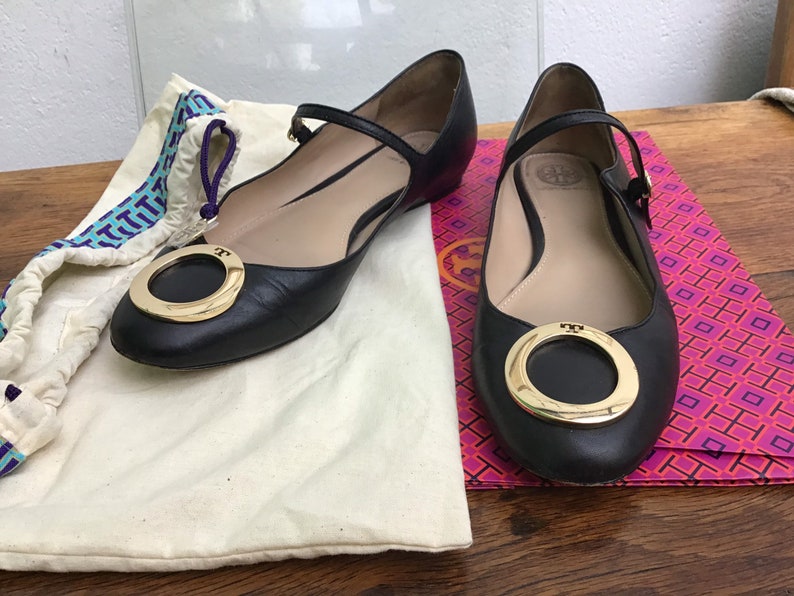 May include: Black leather ballet flats with a gold-tone ring detail on the toe. The flats have a strap across the top of the foot and are displayed with a dust bag and a patterned box.