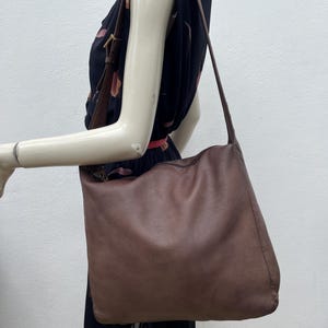 May include: A large, brown leather shoulder bag with a long strap. The bag is worn over the shoulder and hangs down. The leather has a textured appearance. The background is a mannequin wearing a black dress.