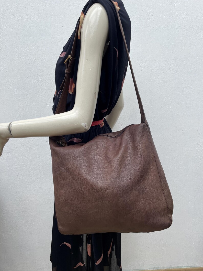 May include: A large, brown leather shoulder bag with a long strap. The bag is worn over the shoulder and hangs down. The leather has a textured appearance. The background is a mannequin wearing a black dress.