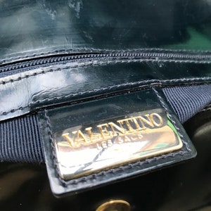 Puede incluir: Un bolso de mano de charol negro con una placa met&aacute;lica rectangular de tono dorado con el texto en relieve VALENTINO LES SACS. El bolso incluye un cierre de cremallera negro, un forro de tela oscura y un bot&oacute;n de presi&oacute;n met&aacute;lico dorado.