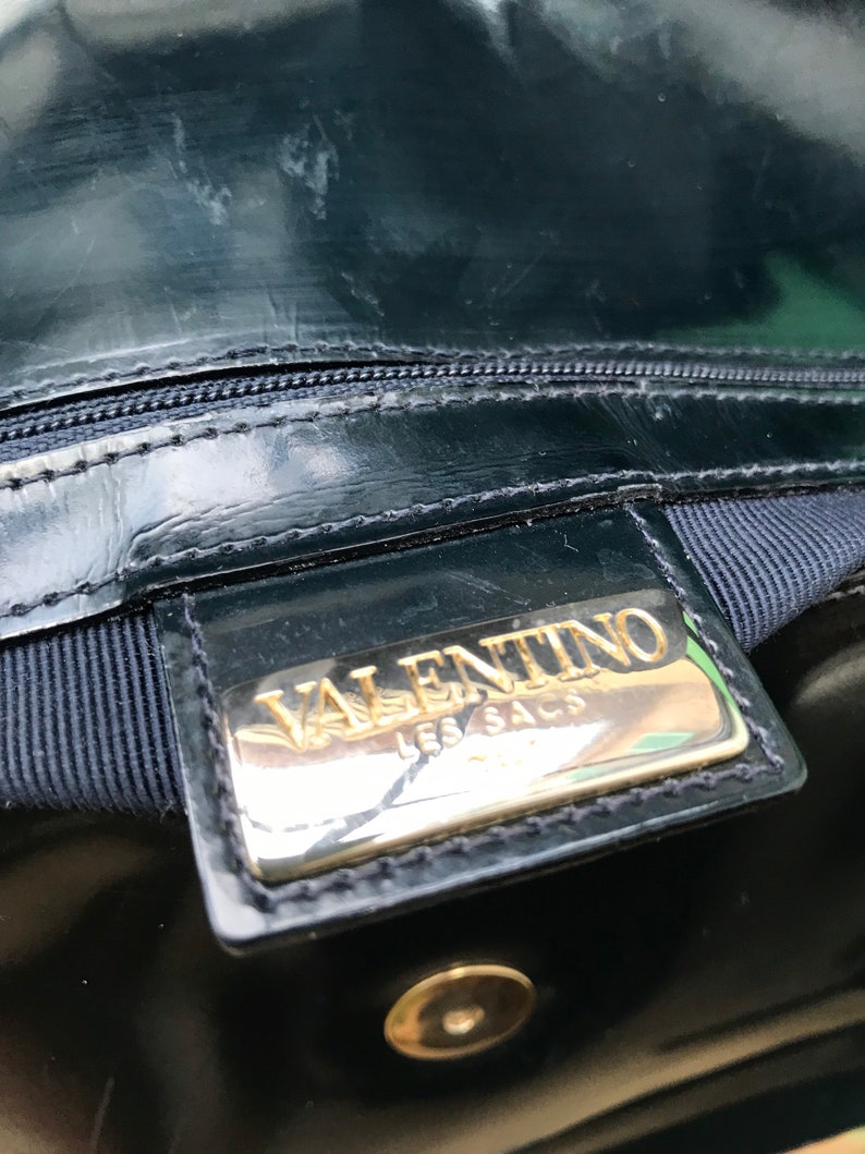 Puede incluir: Un bolso de mano de charol negro con una placa met&aacute;lica rectangular de tono dorado con el texto en relieve VALENTINO LES SACS. El bolso incluye un cierre de cremallera negro, un forro de tela oscura y un bot&oacute;n de presi&oacute;n met&aacute;lico dorado.