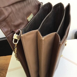May include: A brown leather handbag with multiple compartments. The bag features a gold-tone chain strap and clasp. The interior is a darker brown, and the exterior is a lighter brown. A gold logo is visible on the flap.