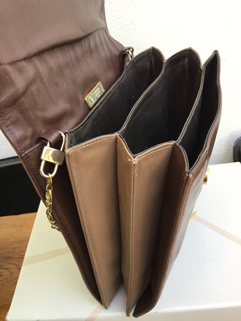 May include: A brown leather handbag with multiple compartments. The bag features a gold-tone chain strap and clasp. The interior is a darker brown, and the exterior is a lighter brown. A gold logo is visible on the flap.