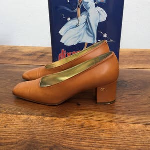 May include: A pair of tan leather pumps with a gold interior and a block heel. The shoes are set against a vintage blue and white advertisement. The heels have a small gold emblem.