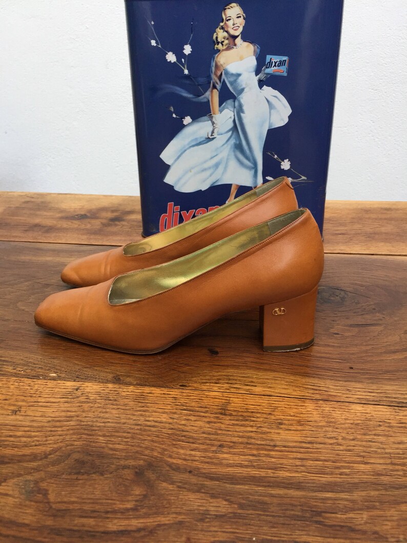 May include: A pair of tan leather pumps with a gold interior and a block heel. The shoes are set against a vintage blue and white advertisement. The heels have a small gold emblem.