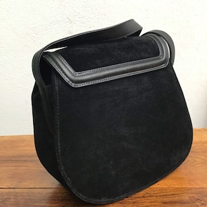 May include: A black suede shoulder bag with a flap closure and a leather strap. The bag has a rounded shape and a contrasting black leather trim. The bag is sitting on a wooden surface.