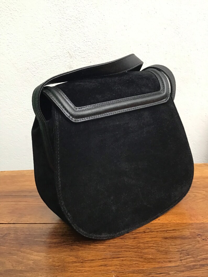 May include: A black suede shoulder bag with a flap closure and a leather strap. The bag has a rounded shape and a contrasting black leather trim. The bag is sitting on a wooden surface.