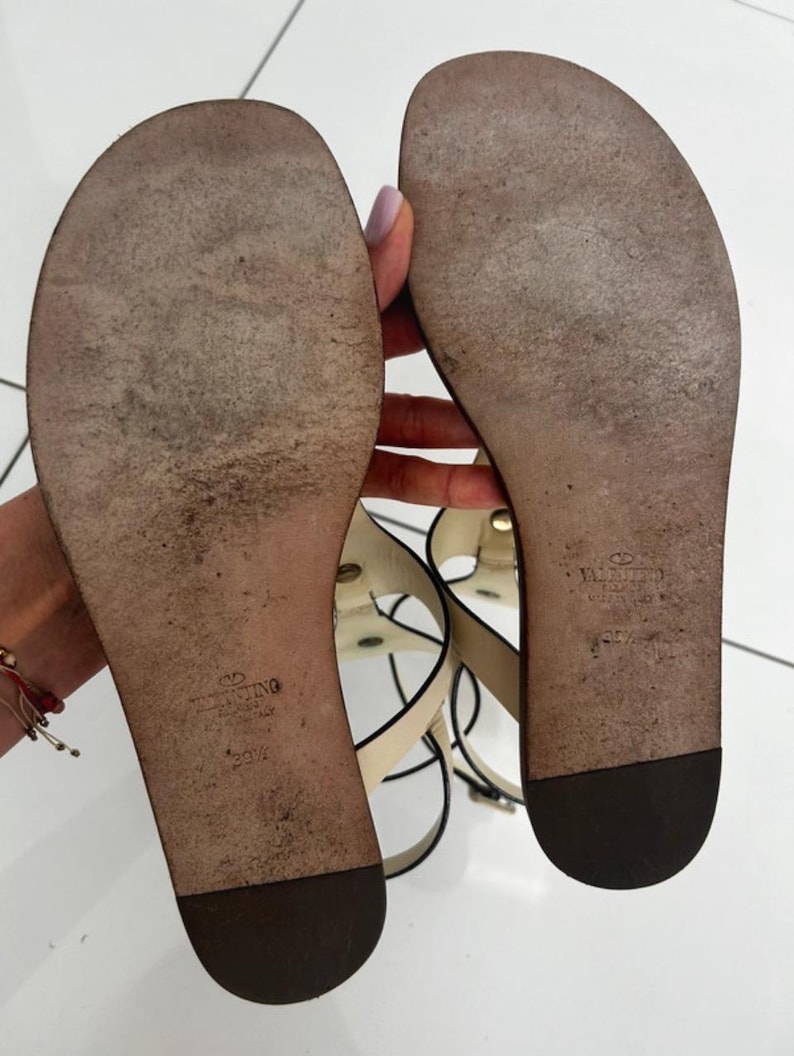 May include: Bottom view of two beige and black leather sandals. The soles are worn and show the Valentino logo and size 39 1/2. The sandals have multiple straps and are on a white surface.