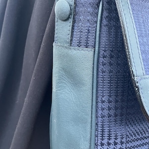 May include: Close-up of a navy blue shoulder bag with a patterned fabric flap and teal leather accents. The bag features a button closure and a leather strap. The background is a dark, solid color.