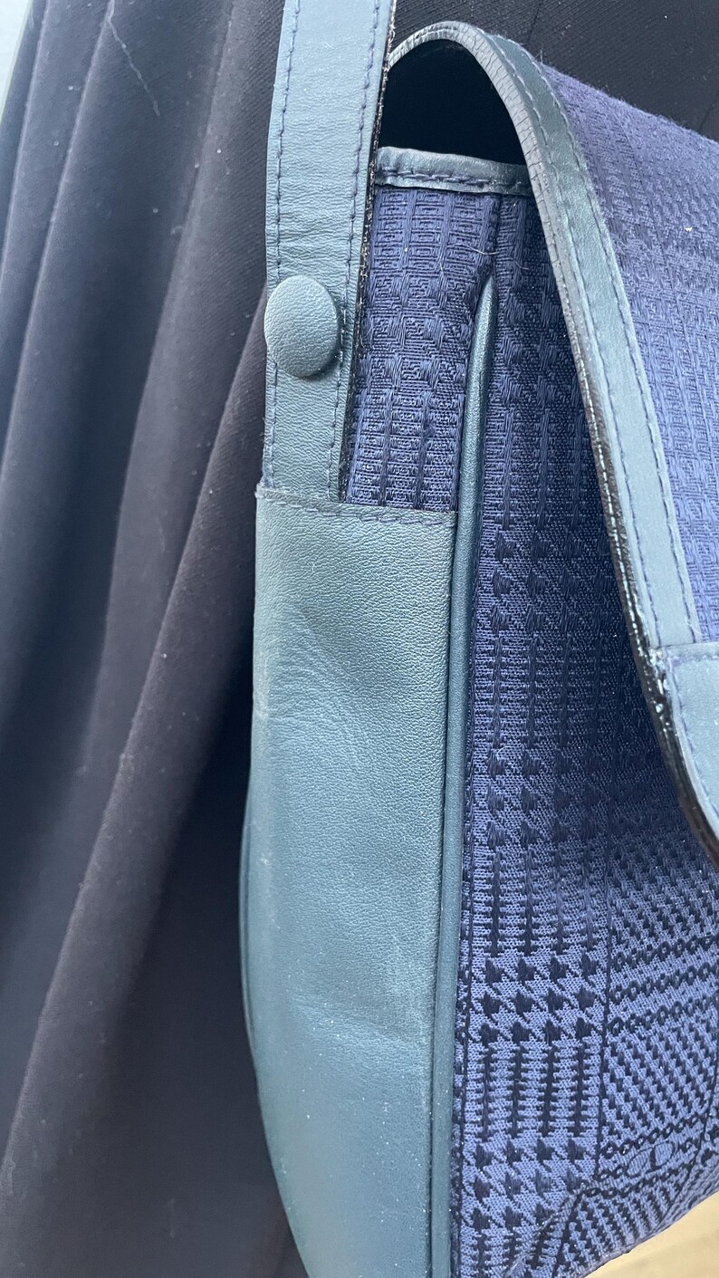 May include: Close-up of a navy blue shoulder bag with a patterned fabric flap and teal leather accents. The bag features a button closure and a leather strap. The background is a dark, solid color.