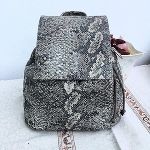 May include: A backpack with a snakeskin pattern in shades of gray and black. The bag has a flap closure and a drawstring opening. The backpack is made of a textured material and has a small handle at the top.
