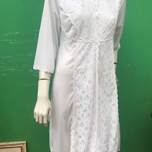 Può includere: Camicia da notte bianca vintage con dettagli in pizzo. La camicia da notte, lunga fino al ginocchio, presenta uno scollo tondo, maniche a tre quarti e un pannello centrale in pizzo intricato. L'abito è esposto su un manichino su sfondo verde.