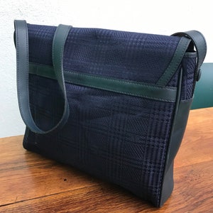 May include: A navy blue messenger bag with a plaid pattern. The bag features a flap closure and a long shoulder strap. The trim and strap are a darker shade of blue. The bag is sitting on a wooden surface.