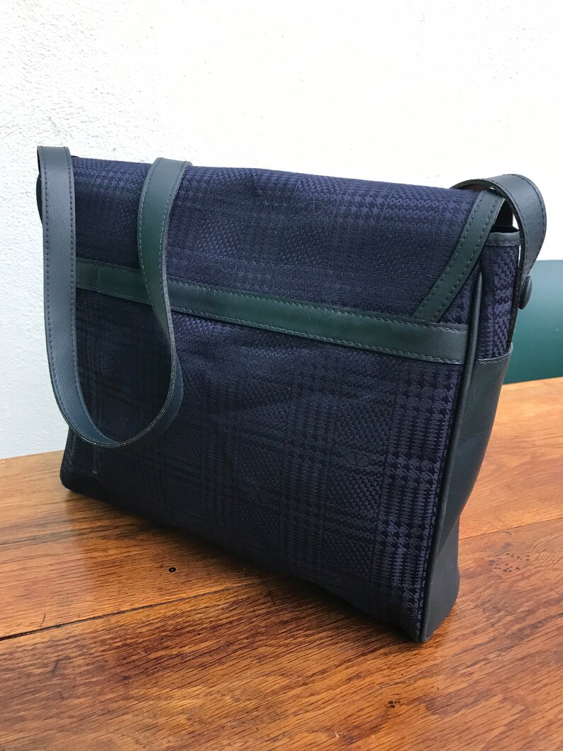 May include: A navy blue messenger bag with a plaid pattern. The bag features a flap closure and a long shoulder strap. The trim and strap are a darker shade of blue. The bag is sitting on a wooden surface.