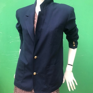 Può includere: Un blazer blu navy con bottoni dorati e collo alla coreana. Il blazer ha due tasche frontali e un piccolo logo sul petto. Il manichino indossa un vestito fantasia sotto.