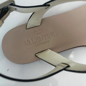 May include: Close-up of a beige leather sandal with thin straps and a silver buckle. The sandal's insole is stamped with the "Valentino Garavani" logo. The straps are black-edged.