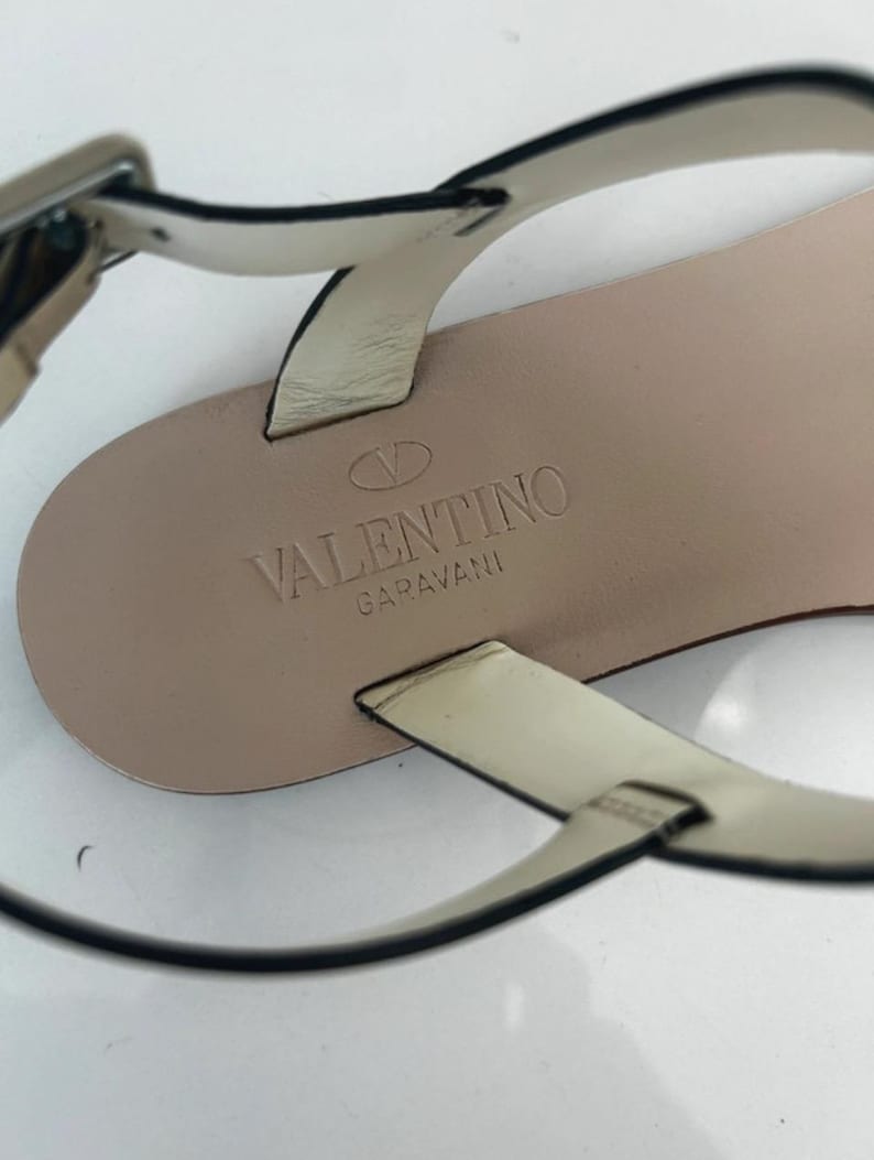 May include: Close-up of a beige leather sandal with thin straps and a silver buckle. The sandal's insole is stamped with the "Valentino Garavani" logo. The straps are black-edged.