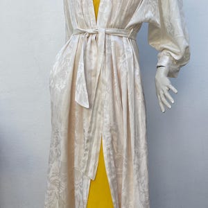 Könnte beinhalten: Langer, cremefarbener Bademantel mit floralem Muster, der mit einem passenden Gürtel in der Taille gebunden wird. Der Bademantel wird über einem leuchtend gelben Kleid getragen. Die Ärmel sind lang und am Handgelenk gerafft.