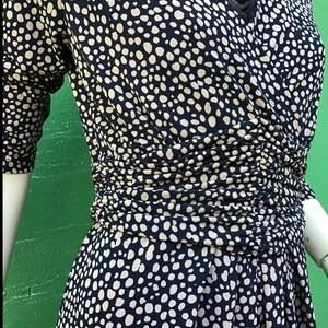 May include: A navy blue dress with a white polka dot pattern. The dress features a V-neckline and three-quarter sleeves. The waist is cinched with gathered fabric, and the skirt flares out. The dress is displayed on a mannequin.