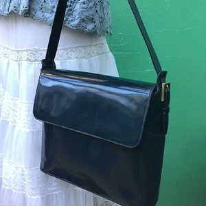 Puede incluir: Un bolso de hombro azul marino oscuro con acabado brillante, que presenta un cierre de solapa rectangular y una correa fina ajustable con herrajes de metal en tono dorado. El bolso tiene una silueta estructurada y una superficie lisa.