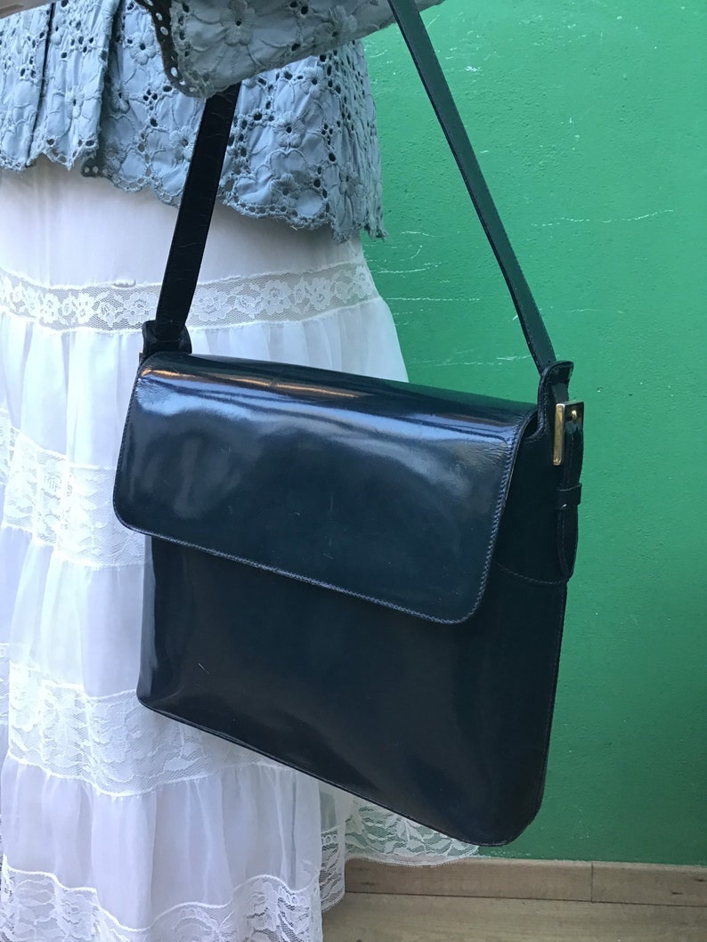 Puede incluir: Un bolso de hombro azul marino oscuro con acabado brillante, que presenta un cierre de solapa rectangular y una correa fina ajustable con herrajes de metal en tono dorado. El bolso tiene una silueta estructurada y una superficie lisa.