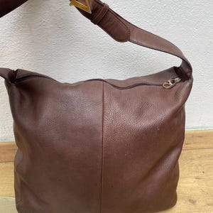 May include: A brown leather shoulder bag with a long strap and a gold-colored buckle. The bag has a zipper closure and a textured surface. The bag is a medium size.