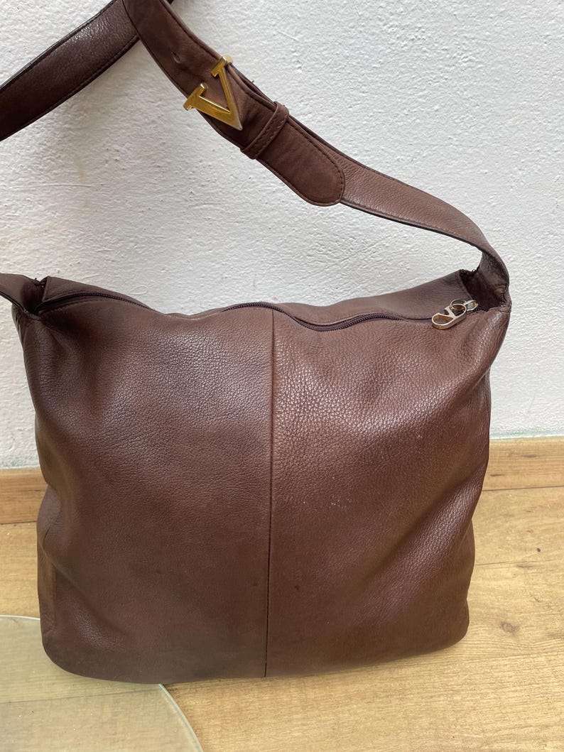 May include: A brown leather shoulder bag with a long strap and a gold-colored buckle. The bag has a zipper closure and a textured surface. The bag is a medium size.