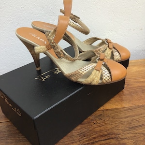 May include: A pair of Prada heeled sandals. The sandals feature a tan leather and woven design with a snakeskin pattern. The heels are wooden and the sandals are on top of a black box.