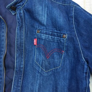 Peut inclure: Une veste en jean bleu avec un logo Levi's rouge sur la poche. La veste a une fermeture &eacute;clair et un col montant.
