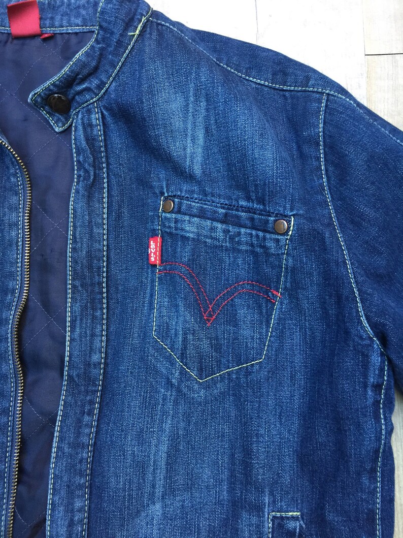 Peut inclure: Une veste en jean bleu avec un logo Levi's rouge sur la poche. La veste a une fermeture &eacute;clair et un col montant.