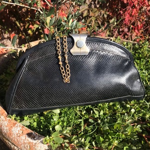 Op de afbeelding: Een zwarte clutch van slangenleer met een gouden kettingriem en een zilveren sluiting. De clutch ligt op een bed van groene planten.